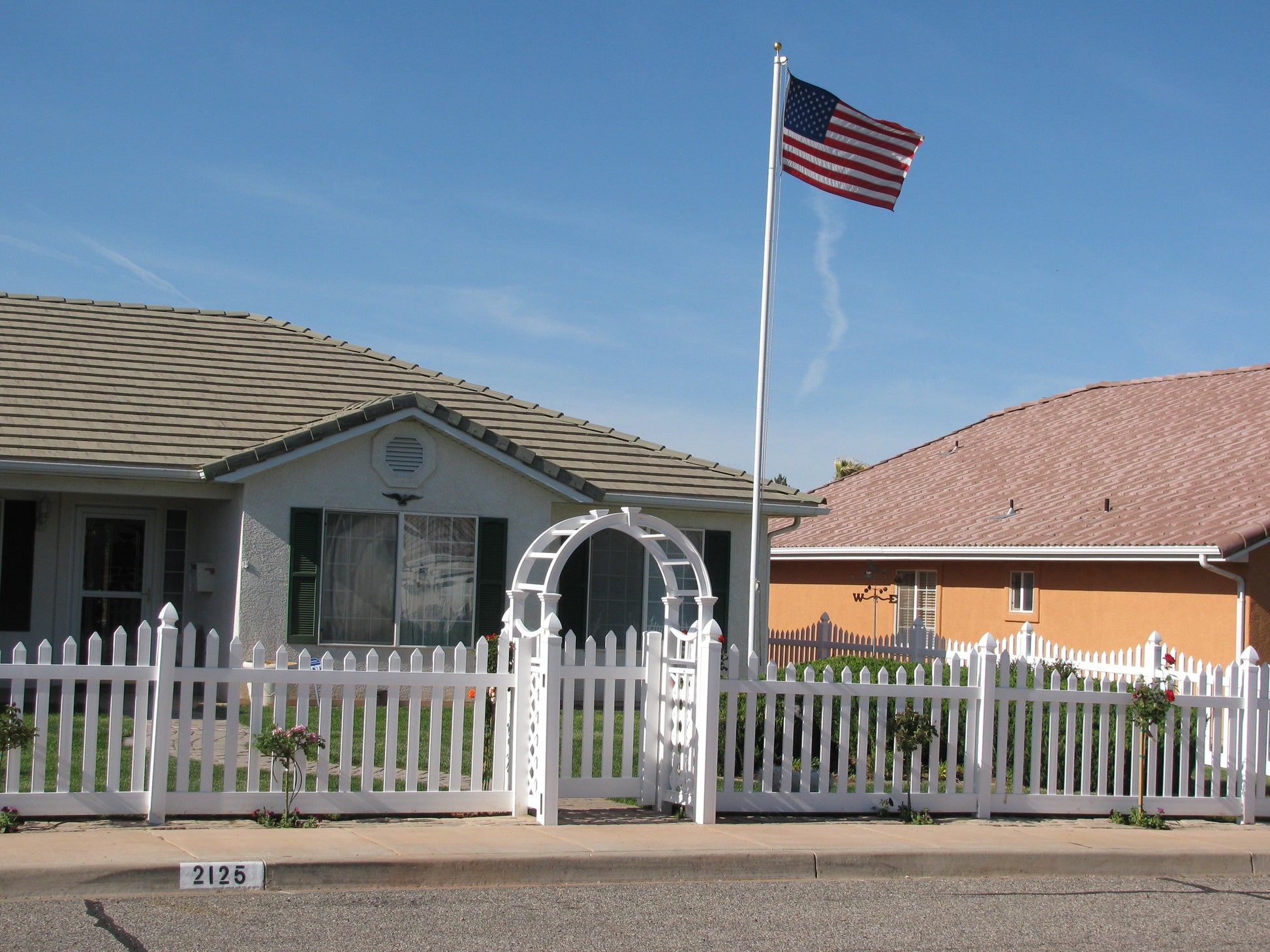 Weatherables Hampshire white vinyl picket fence in front of a house with an American flag on a clear day.