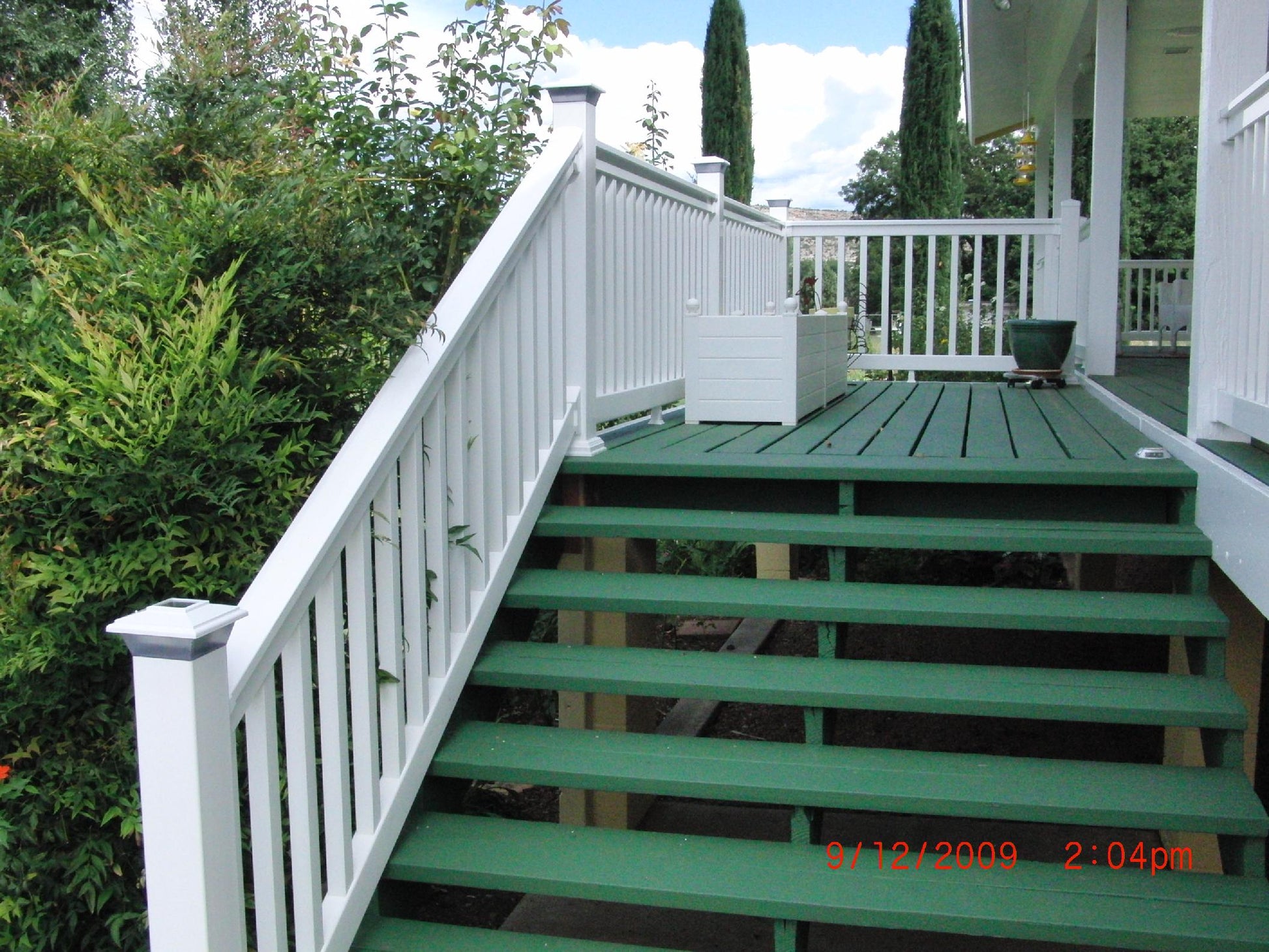 Green wooden steps leading to a  deck with Weatherables Washington white vinyl deck railing, surrounded by greenery.