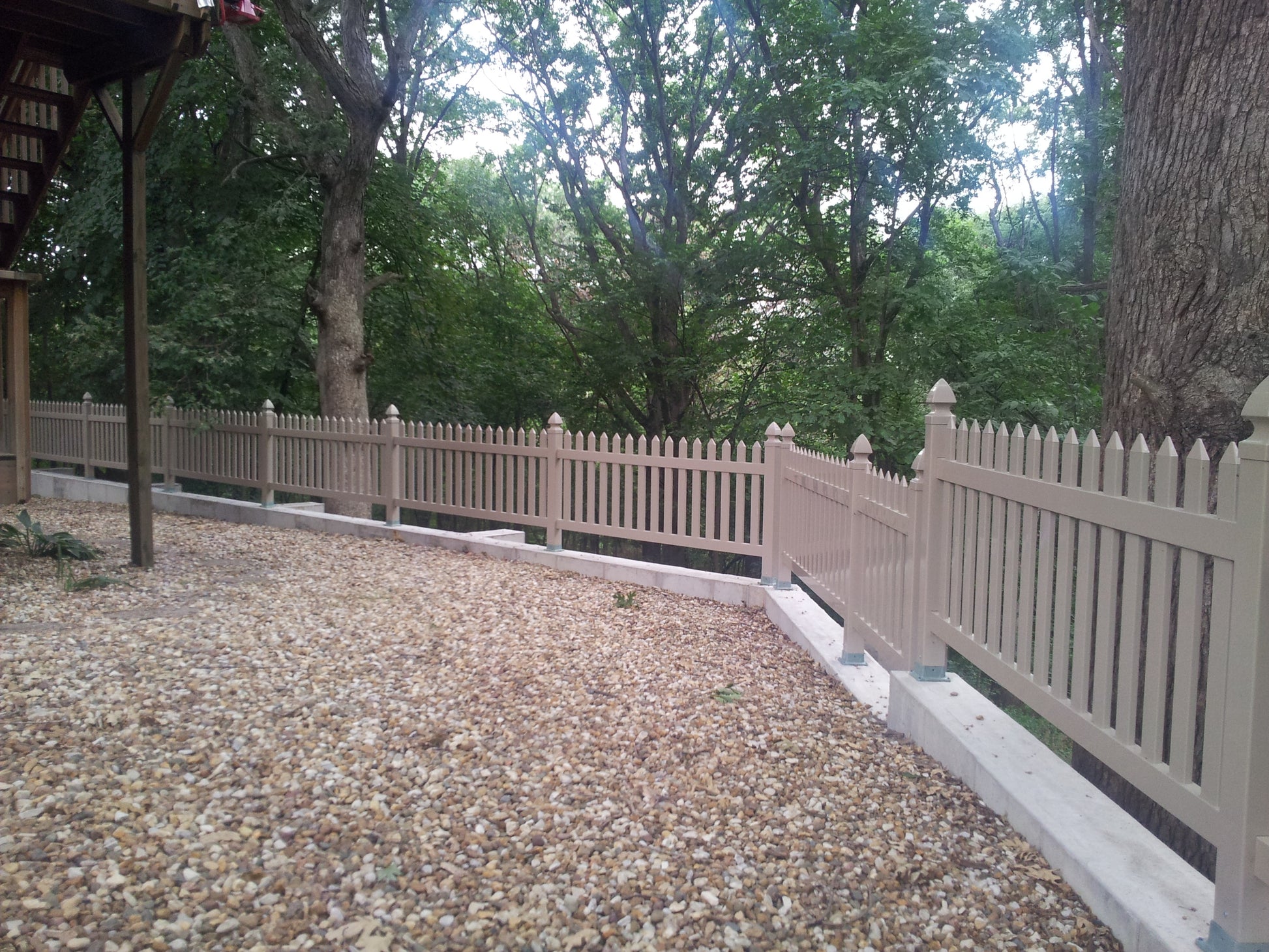 Paved area with a Weatherables Chelsea khaki vinyl picket fence and trees in the background