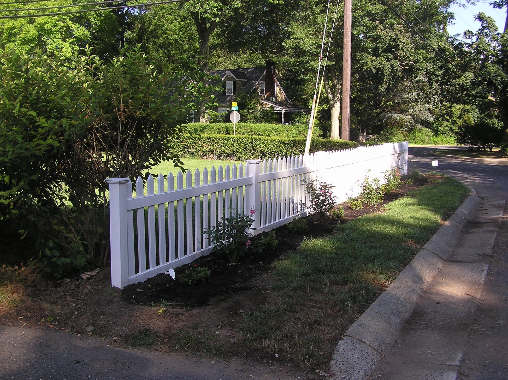 Weatherables Chelsea white vinyl picket fence along a residential street with greenery and a house in the background.