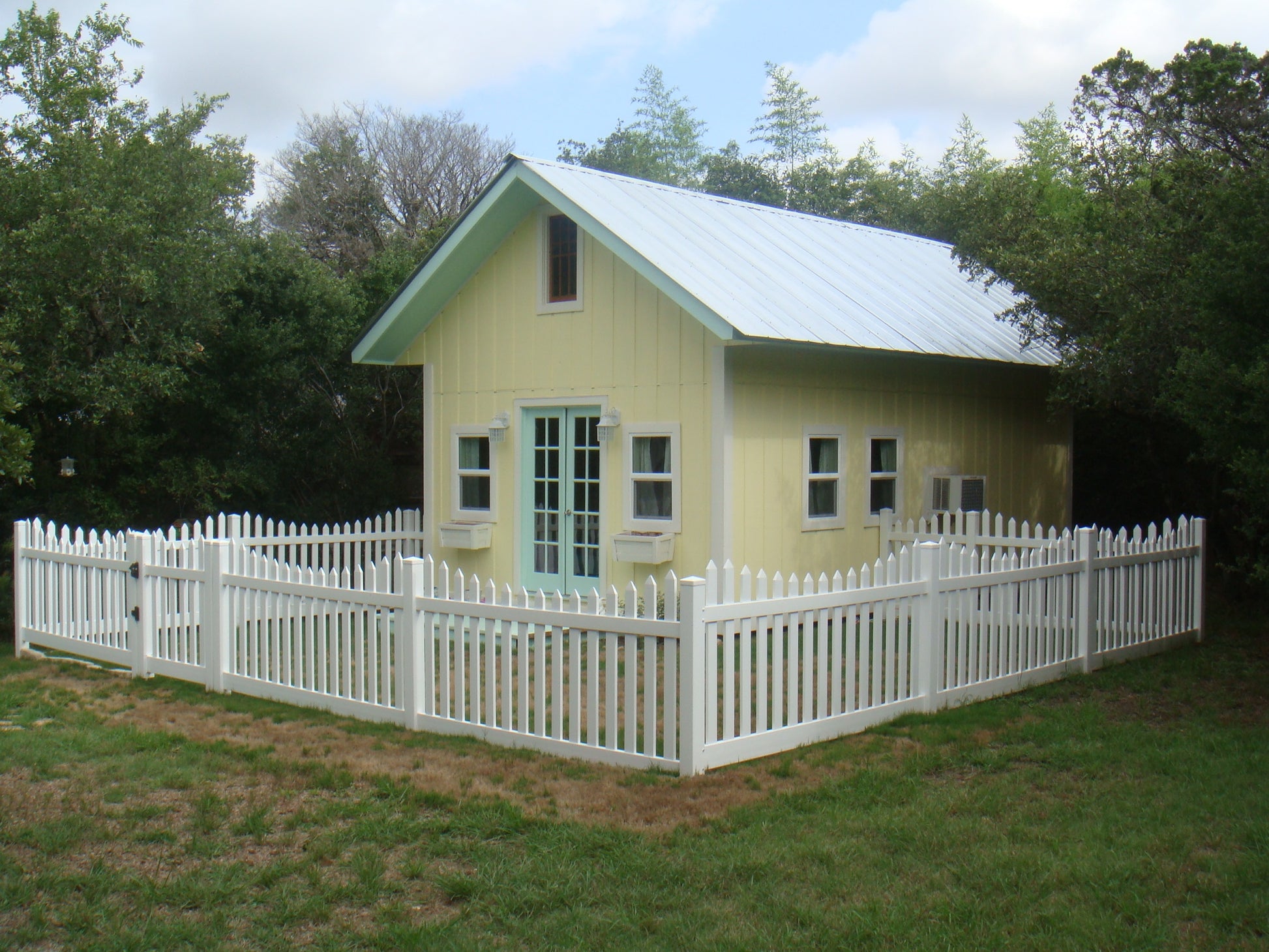 Small yellow house with a Weatherables Hampshire white vinyl picket fence surrounded by trees