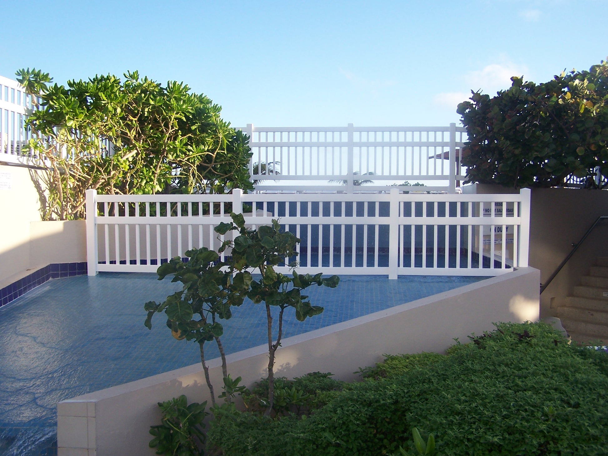 Pool area with a Weatherables Neptune white vinyl pool fence, trees, and clear blue sky