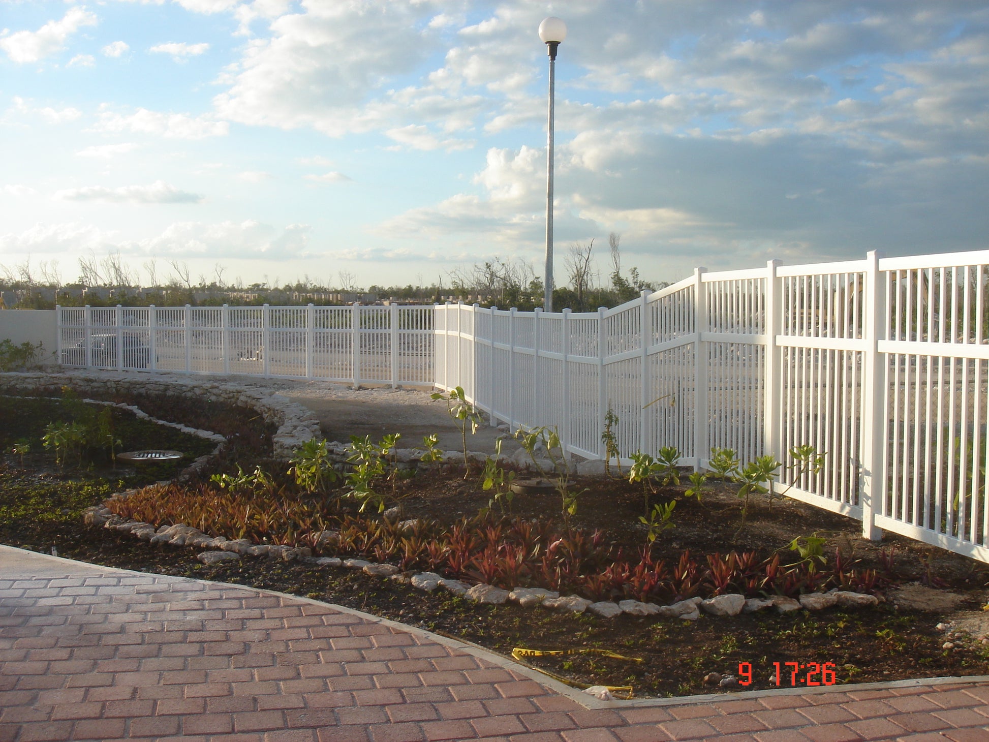 Weatherables Neptune white vinyl pool fence with garden bed and paved walkway under a cloudy sky.