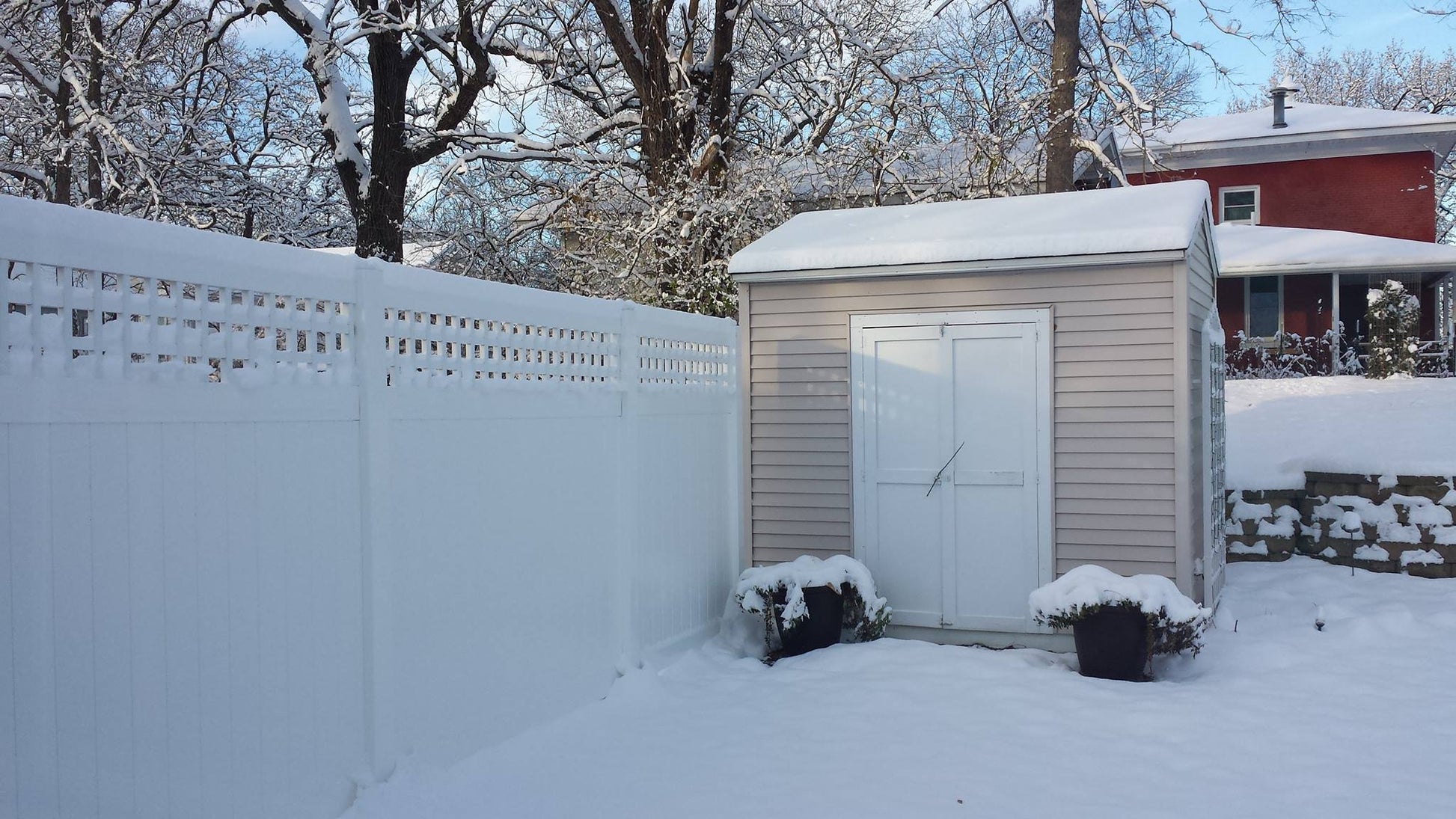 Small shed covered in snow next to a Weatherables Scottsdale white vinyl privacy fence with trees and a house in the background.