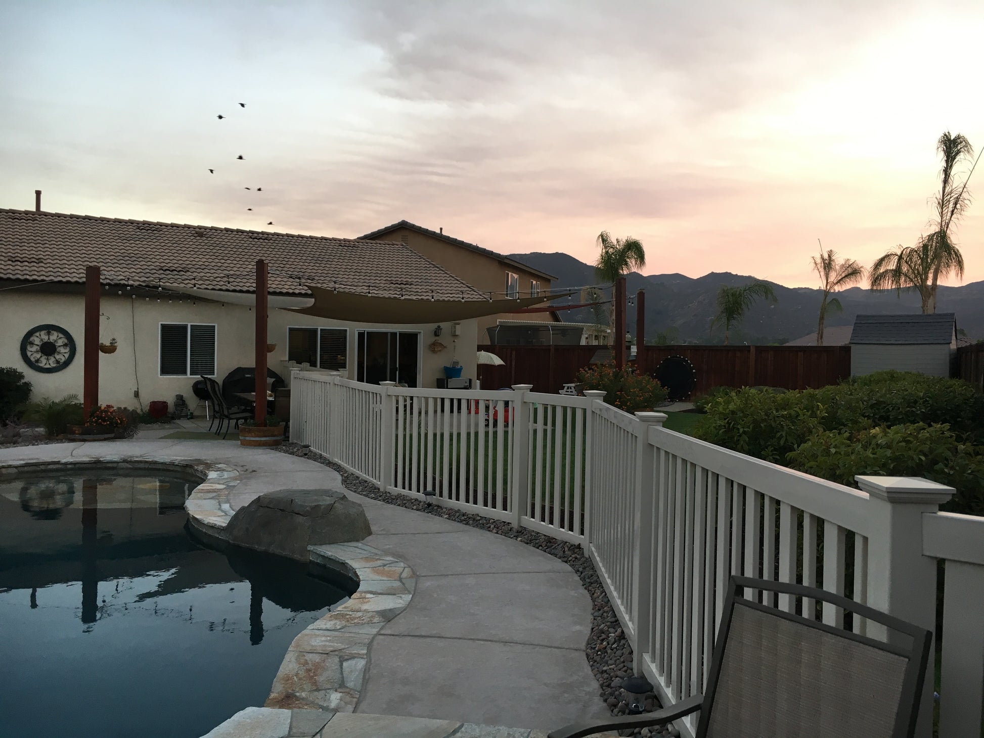 Pool area with a Weatherables Atlantis tan vinyl pool fence, chairs, and a house in the background during sunset.
