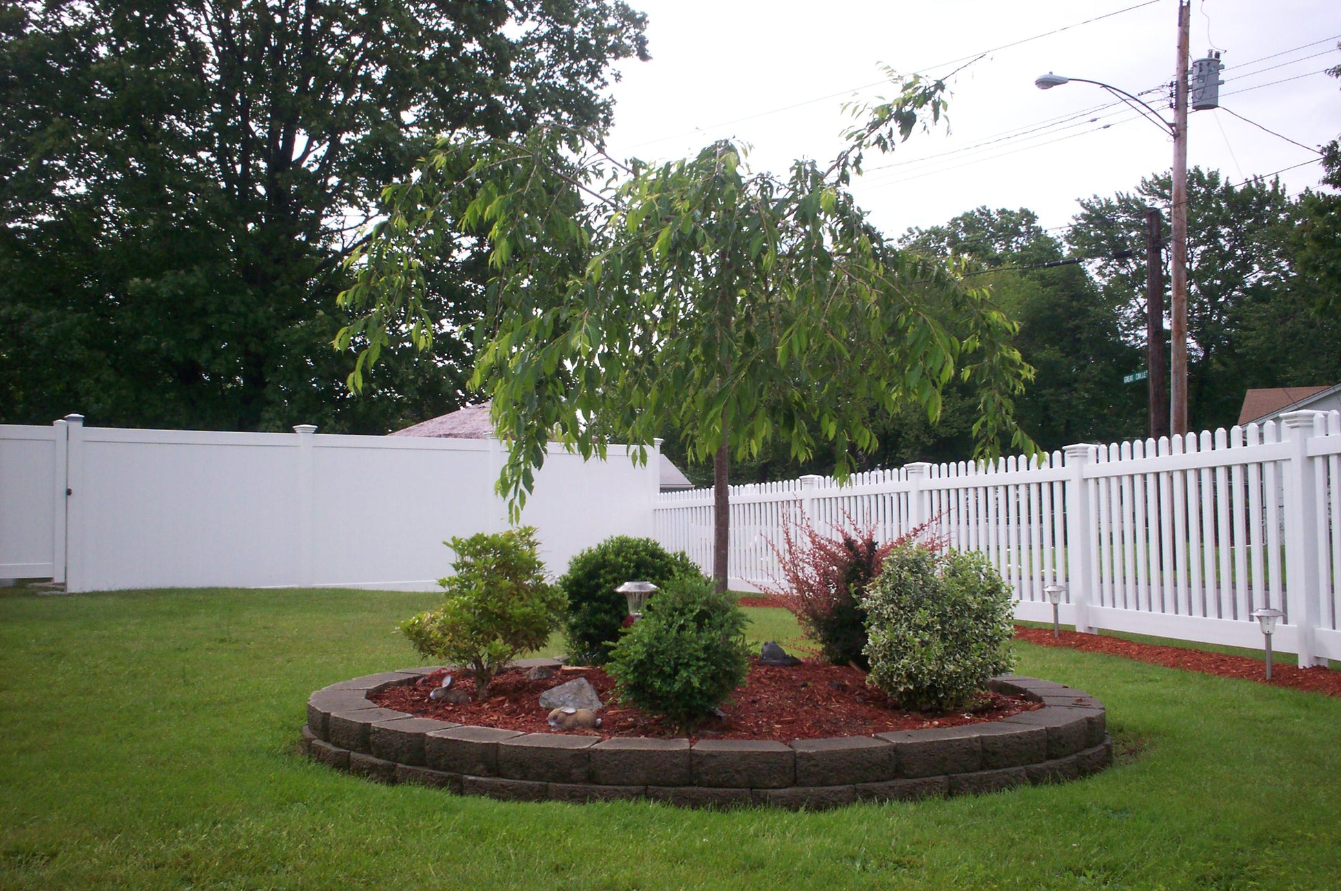 Small garden with plants and a tree in a backyard setting surrounded by a Weatherables Plymouth white vinyl picket fence