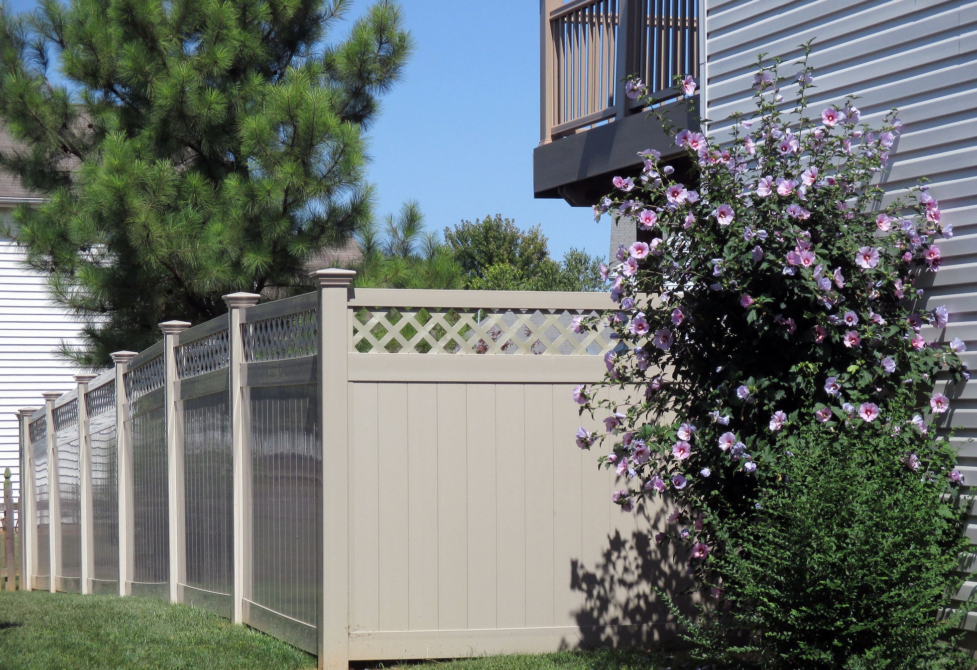 Weatherables Ashton khaki vinyl privacy fence with lattice design, greenery, and a flowering bush in front of a house.