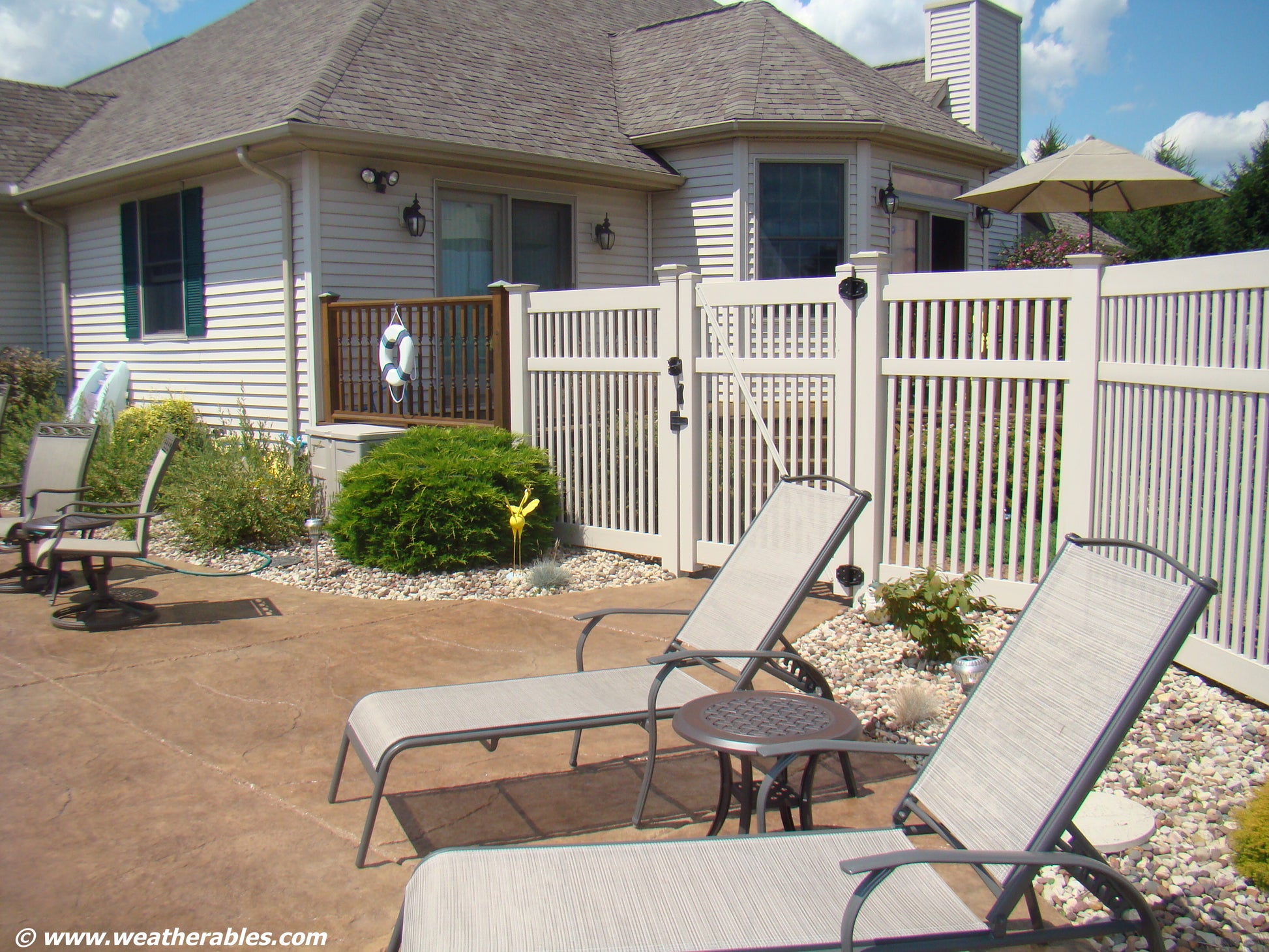 Outdoor patio area with lounge chairs, table, and Weatherables Neptune tan vinyl 5-foot pool fence and house in the background