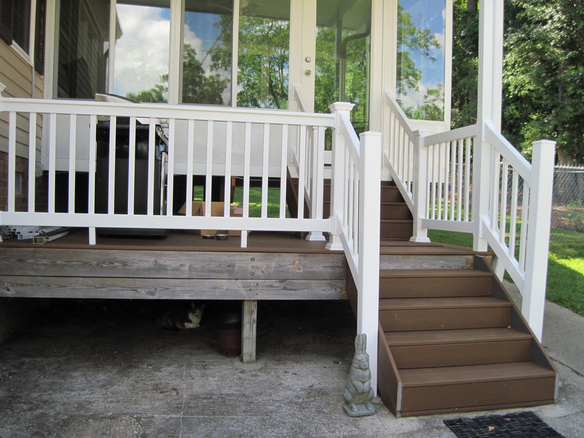Wooden deck with Weatherables Yokrshire white vinyl deck railing and brown steps leading to a house. Underneath the deck is a cat, hiding in darkness for fear it would appear ugly next to the railing.