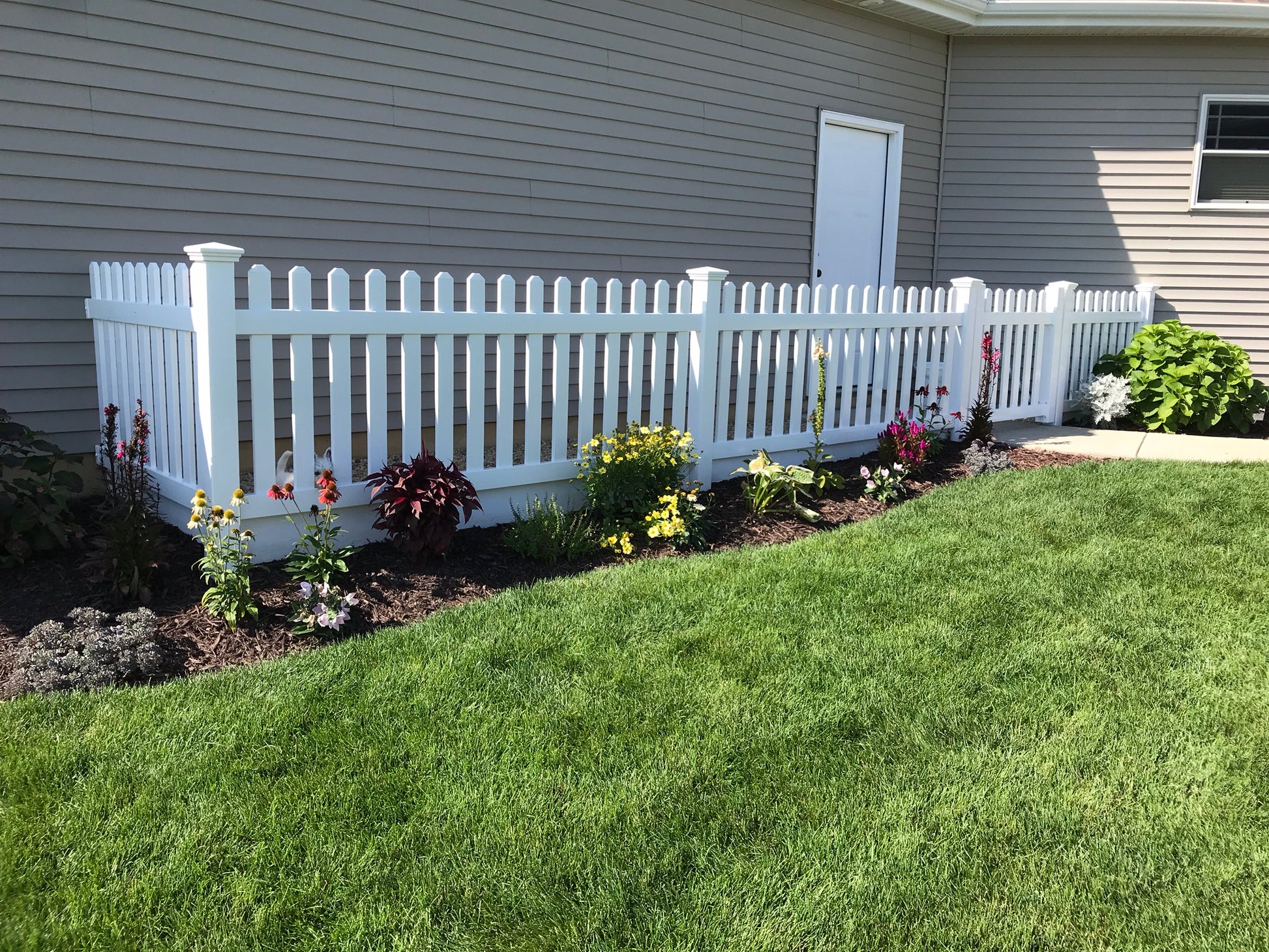 Lawn with a Weatherables Chelsea white vinyl picket fence and garden bed in front of a house.