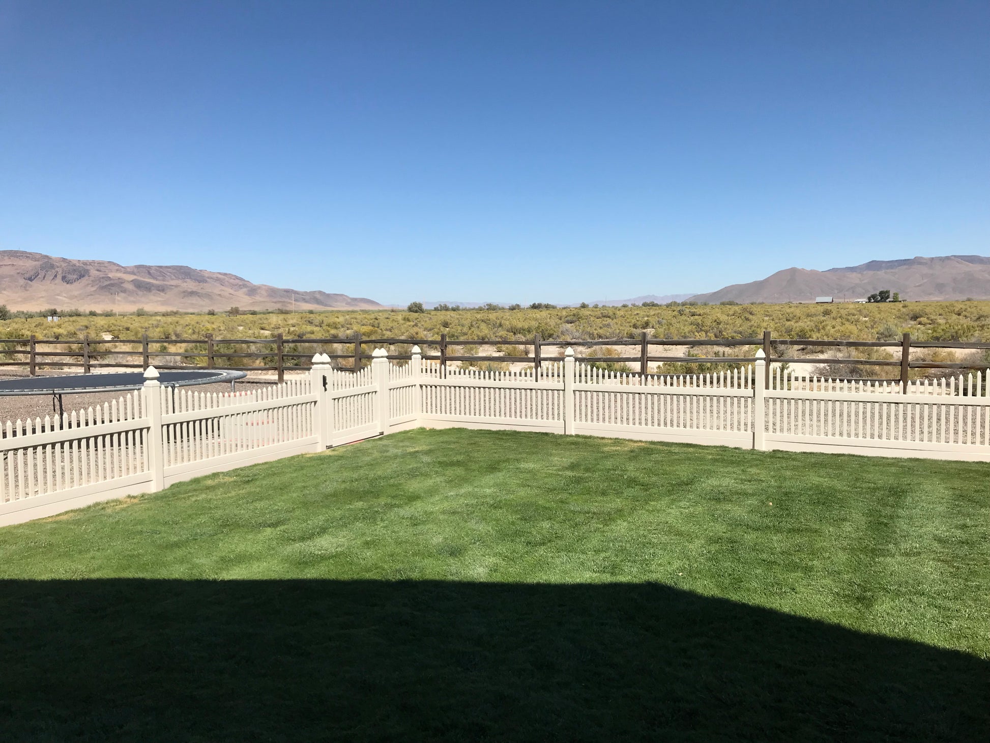 Backyard with a Weatherables Barrington tan vinyl picket fence, green grass, and mountains in the distance under a clear blue sky.