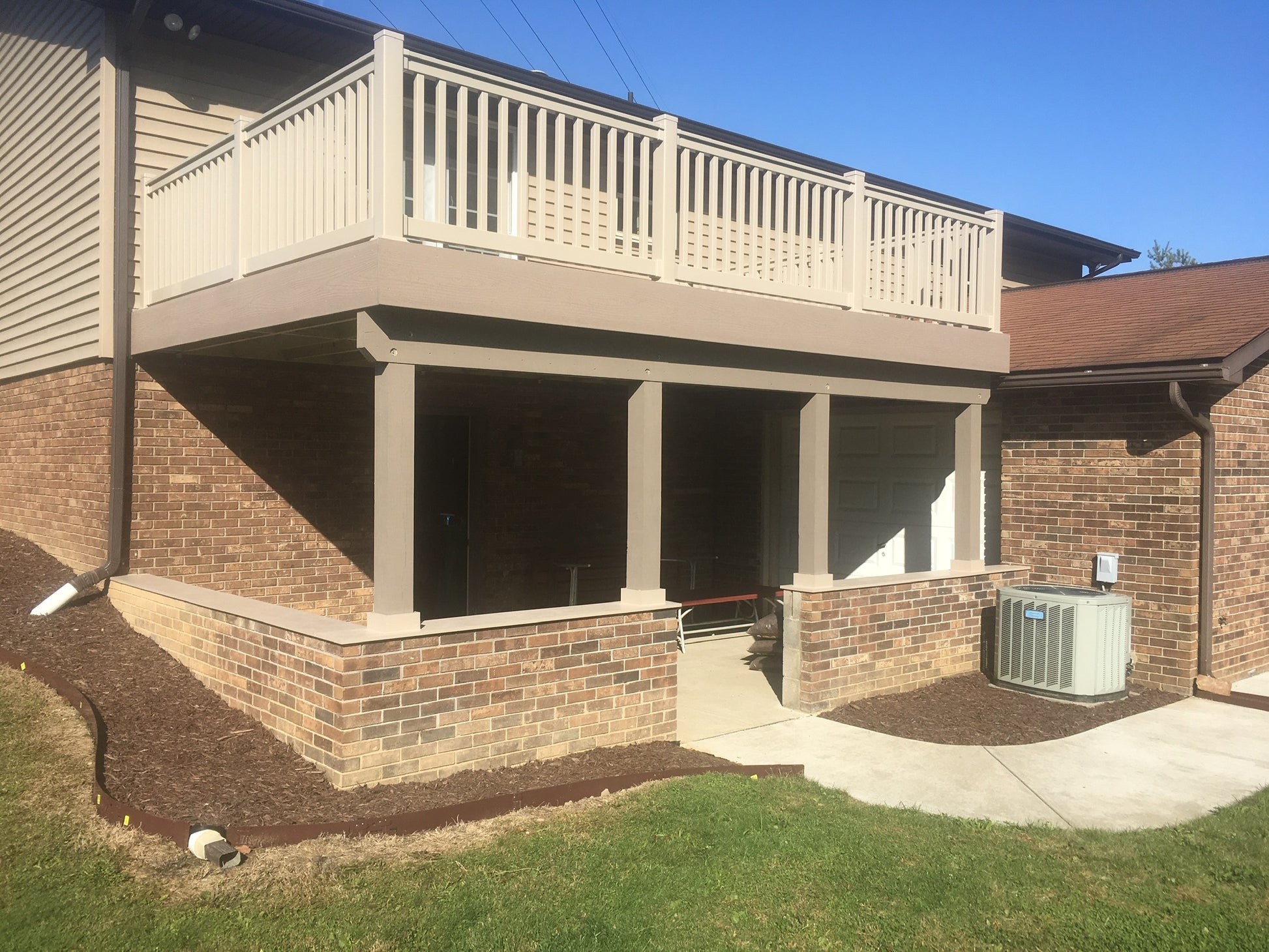Brick house with a Weatherables Berkshire tan vinyl deck railing surrounded porch and air conditioning unit on a clear day 