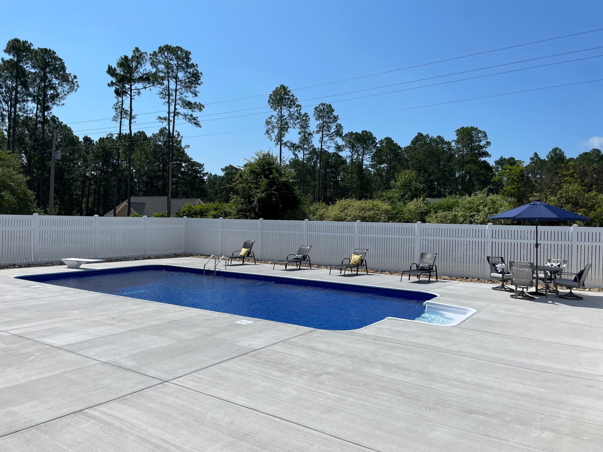 In-ground pool with lounge chairs and a Weatherables Huntington white vinyl semi-privacy fence, surrounded by trees and a clear blue sky.