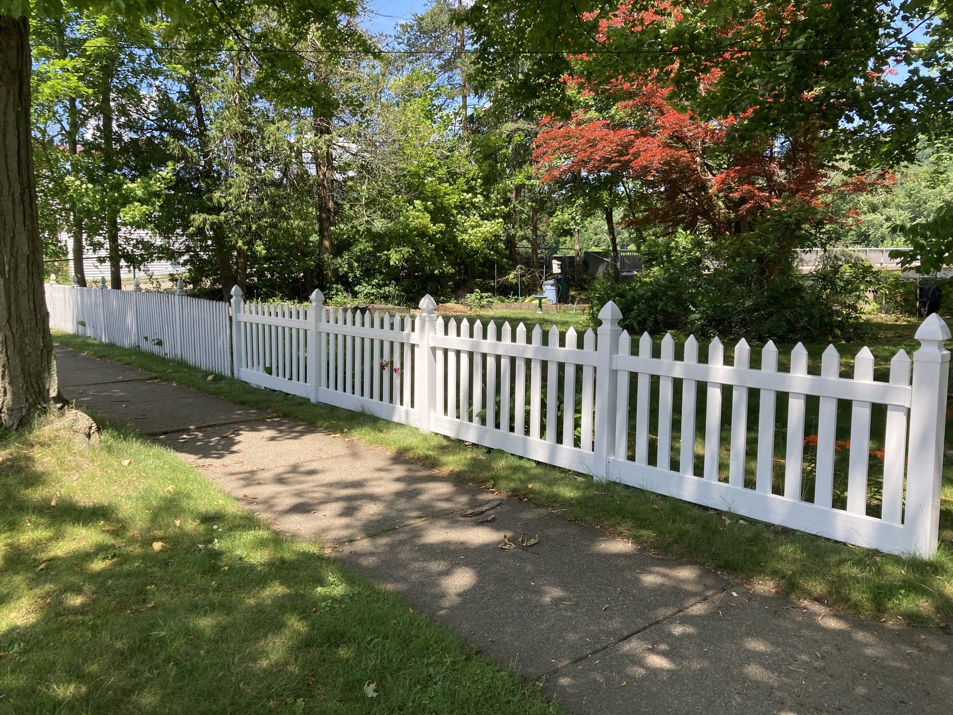 Weatherables Plymouth white vinyl picket fence along a sidewalk with trees and grass in the background