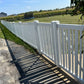 Weatherables Sarasota white vinyl pool fence along a sidewalk with a view of a field and trees in the background.
