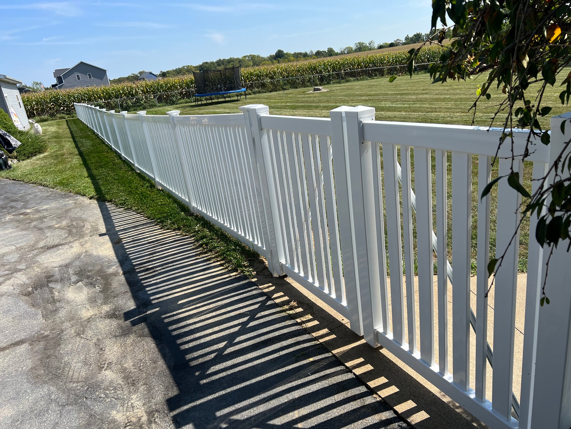 Weatherables Sarasota white vinyl pool fence along a sidewalk with a view of a field and trees in the background.
