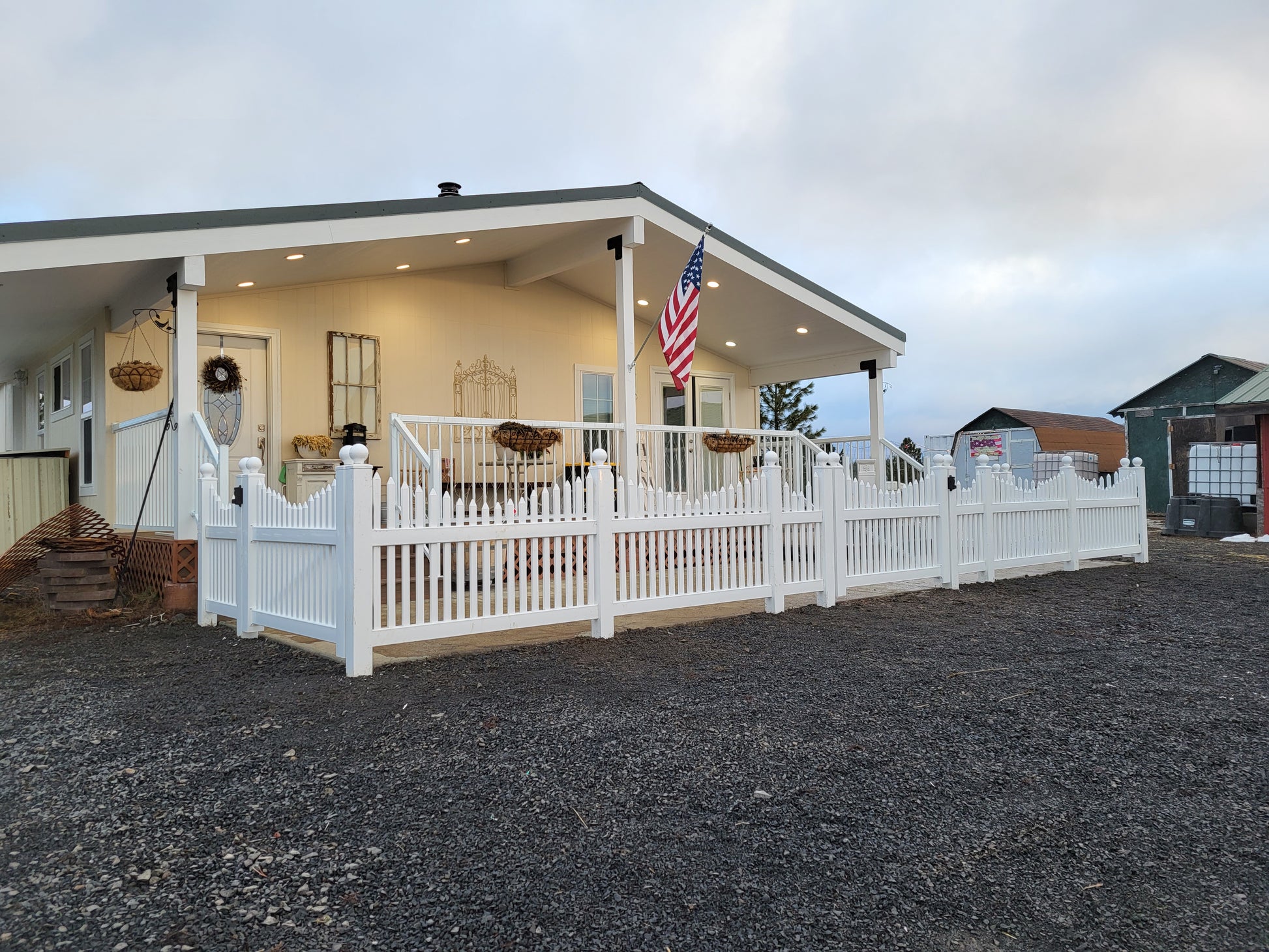 White house with a porch and American flag, surrounded by gravel, Weatherables Barrington white vinyl scalloped picket fence and another building in the background.