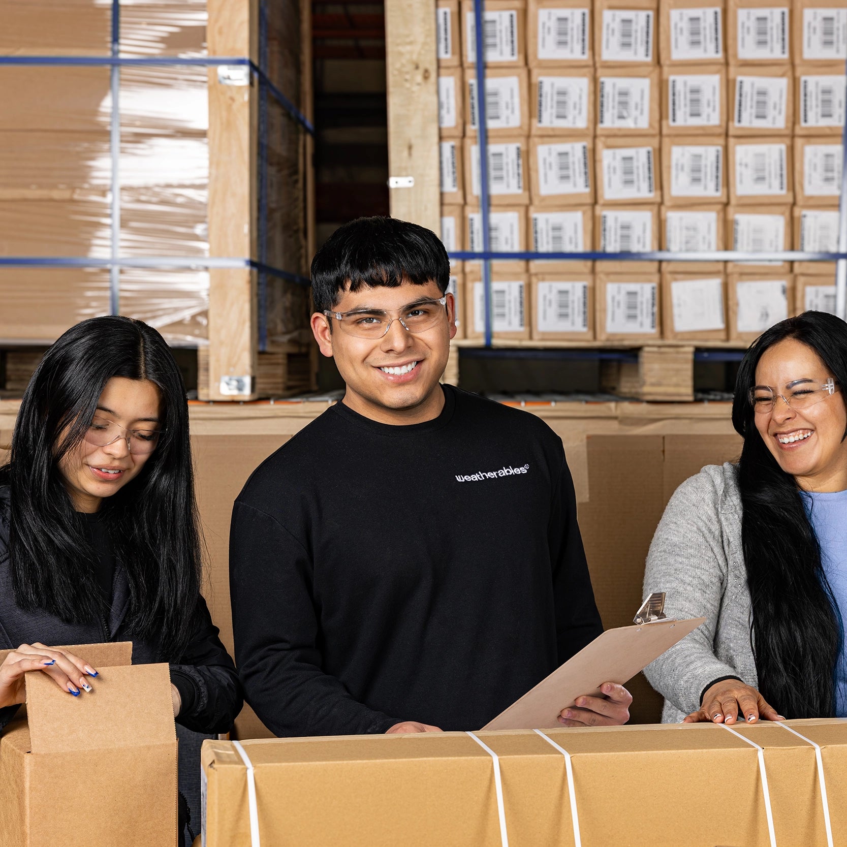 Three people in a warehouse setting with boxes and a person wearing a black shirt with visible text.
