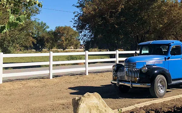 Vintage blue truck parked near a white fence with trees in the background