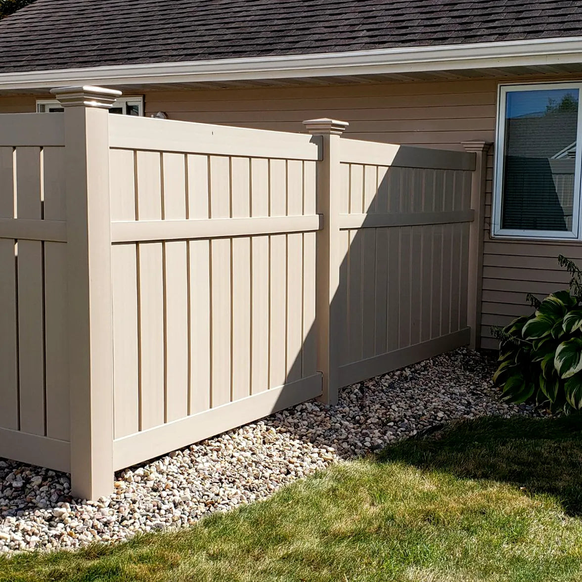 Beige wooden fence next to a house with a window, grass, and gravel.