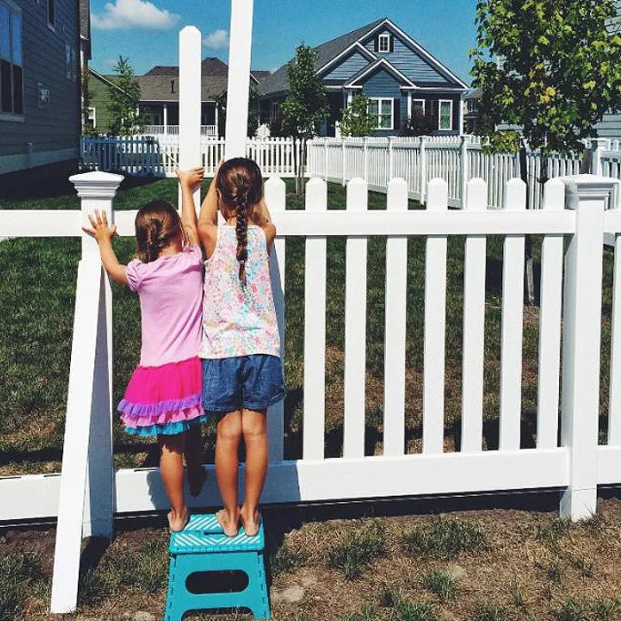 Two children standing on a blue step stool against a white picket fence with a house in the background.