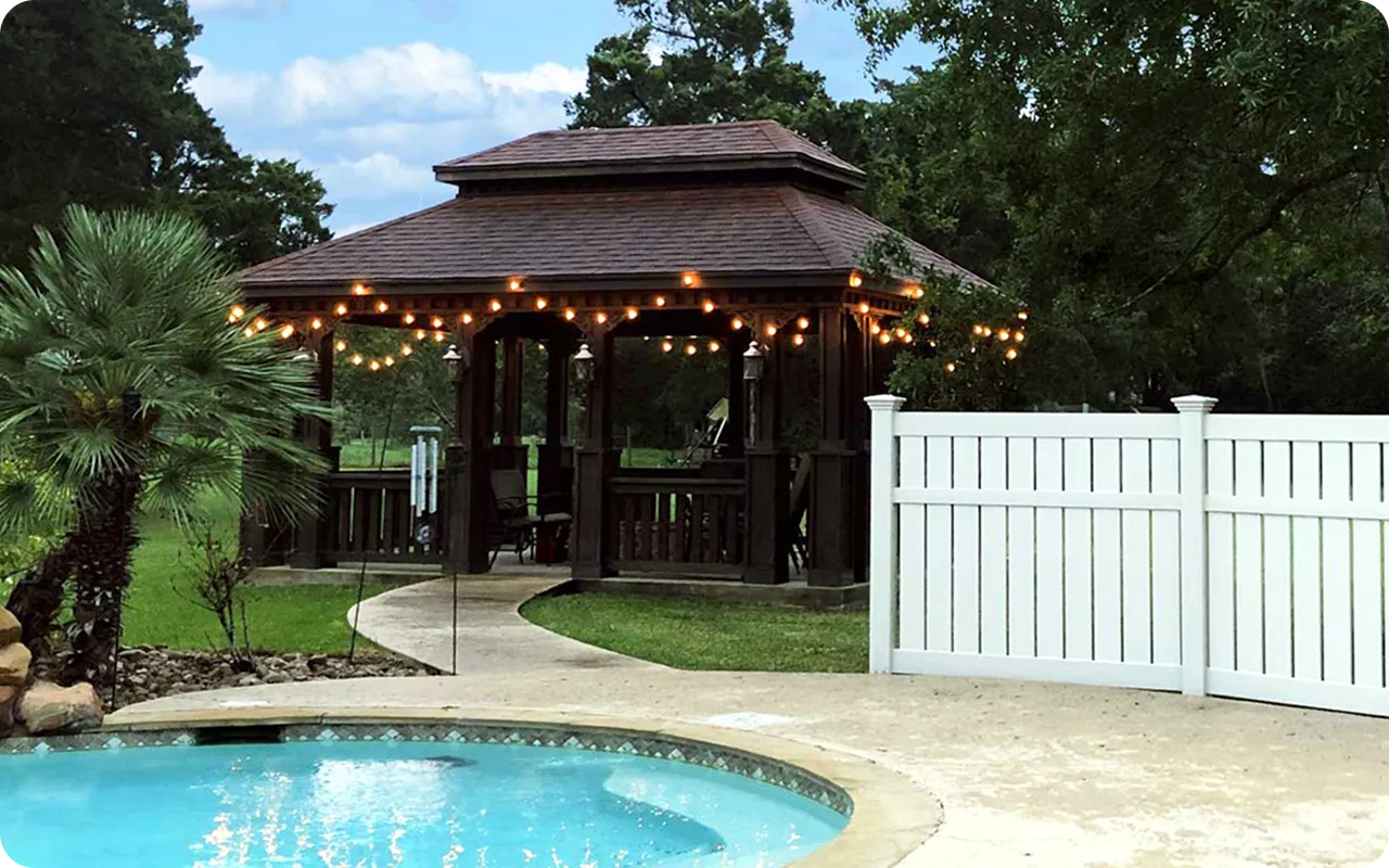 Wooden gazebo with string lights by a poolside with a white fence and trees in the background.